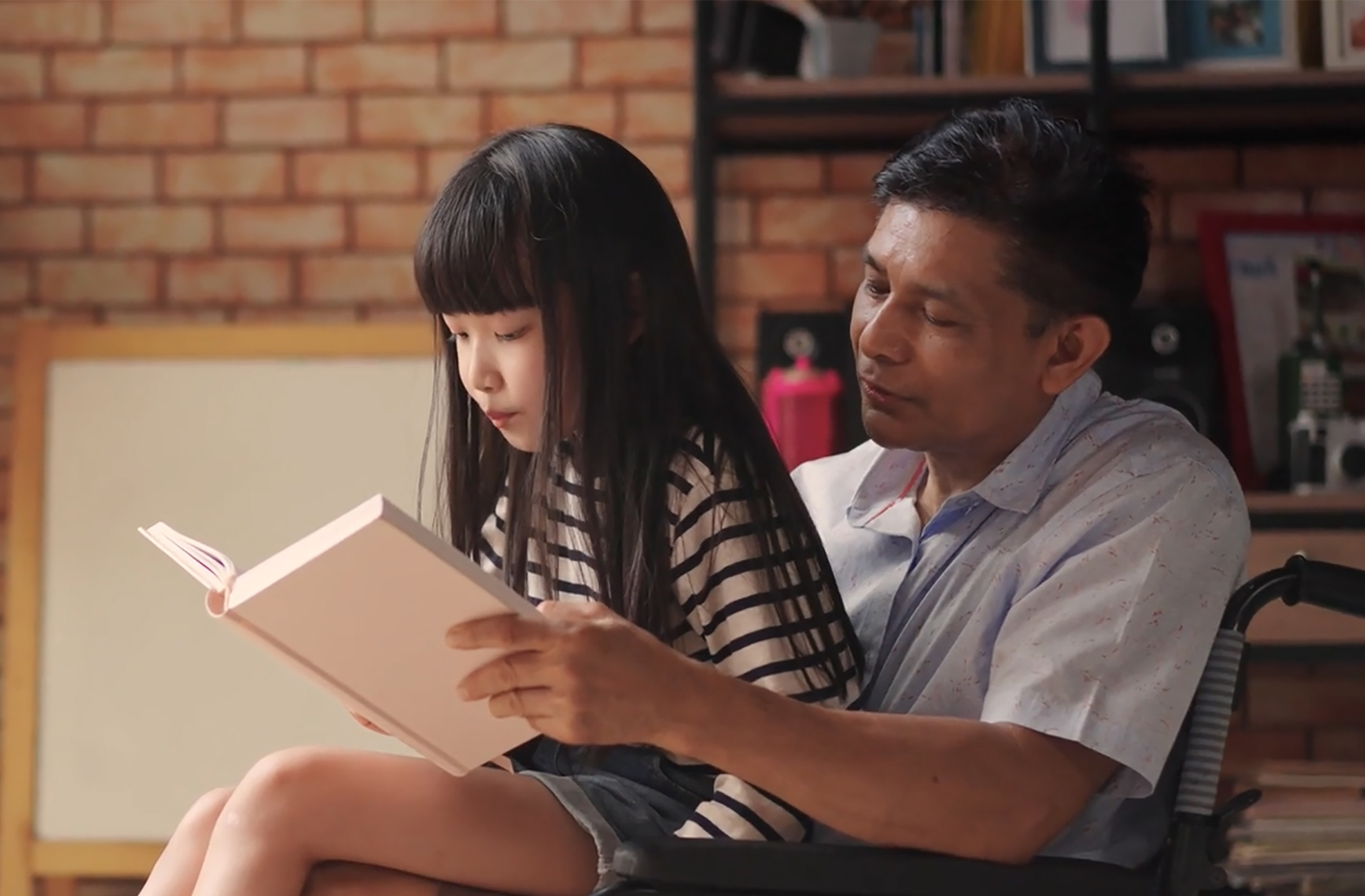 Un père en fauteuil roulant et sa fille assise sur ses genoux lisent un livre ensemble.
