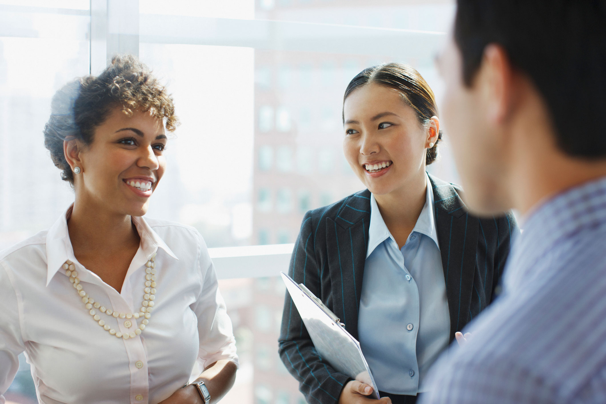 Two businesswomen smile and chat, one holding a clipboard, while a colleague listens in a sunlit office.