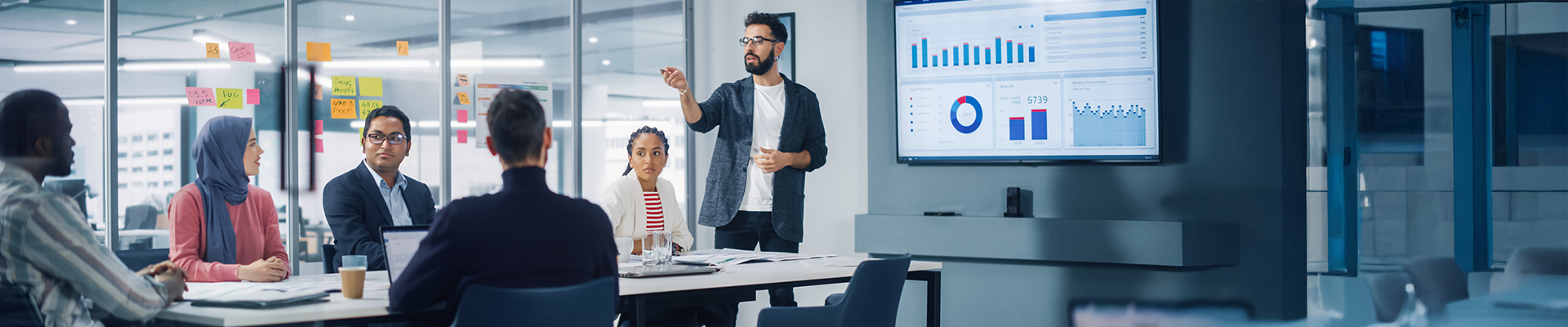Diverse group of professionals in a modern office boardroom as one man presents data on a large screen displaying charts and graphs.