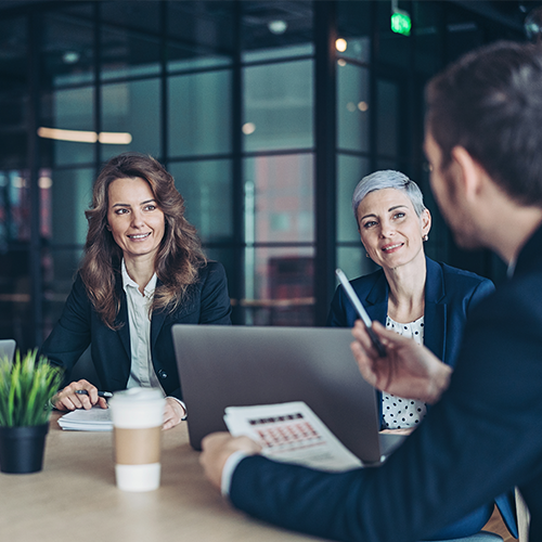 Three business professionals in a modern glass-walled office having a discussion around a laptop, with documents and coffee cups on the table.