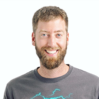 Smiling man with a light brown beard and short hair, wearing a gray graphic T-shirt, posed against a plain white background.