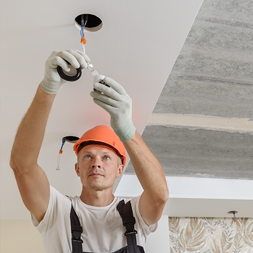 Electrician wearing gloves and an orange hard hat installs ceiling lighting in a building under renovation, holding wiring and electrical tape.