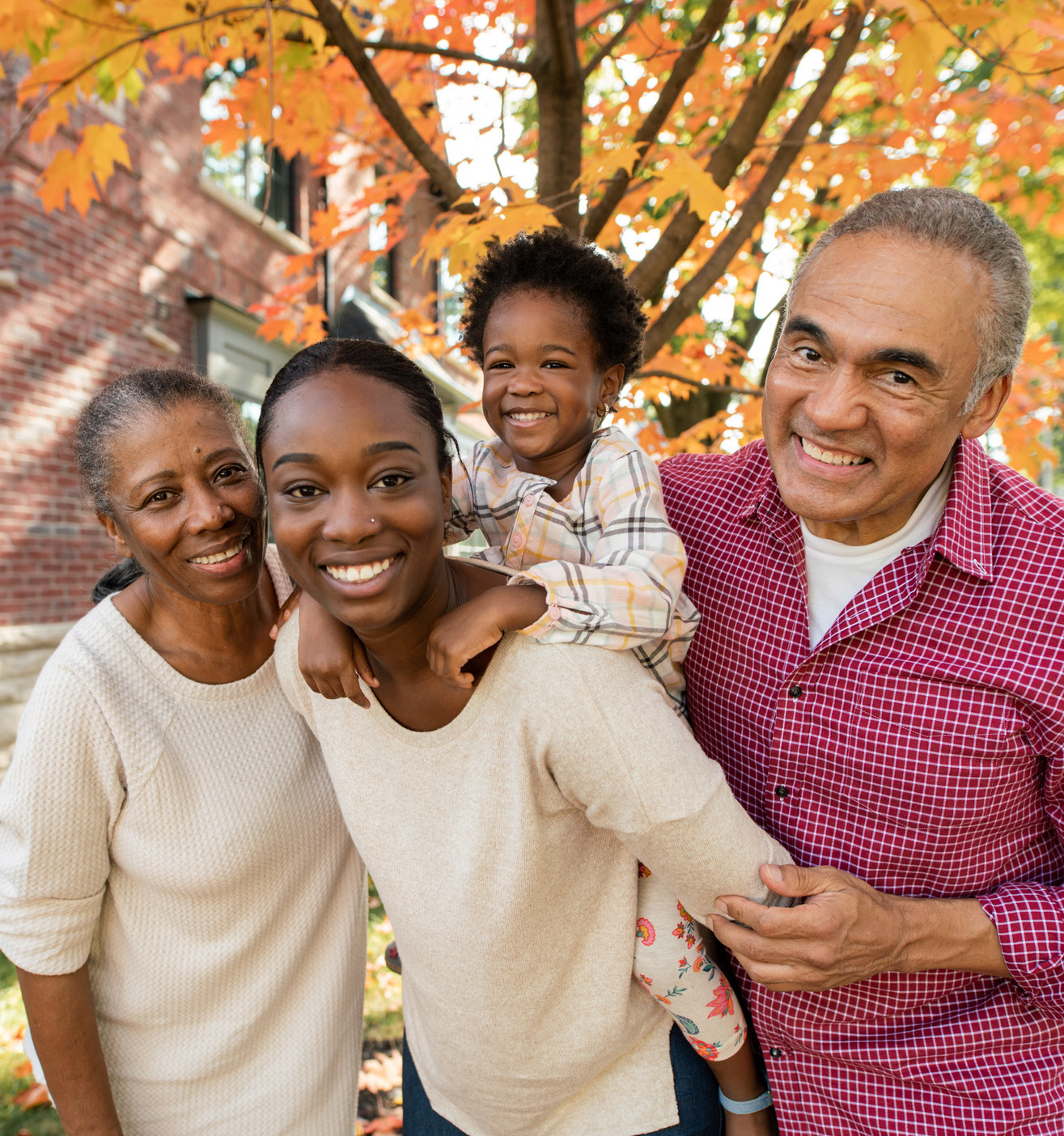 Smiling multigenerational family of four posing together outdoors in front of autumn-coloured trees and a brick building.