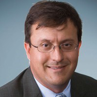 Professional headshot of a man with short brown hair and glasses, wearing a dark suit jacket and light blue shirt, smiling slightly against a muted blue background.