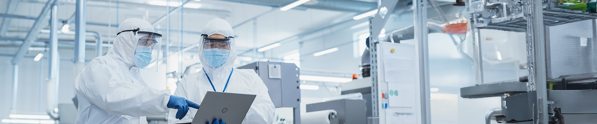 Two scientists in full cleanroom suits, gloves, face shields, and masks reviewing data on a laptop inside a sterile pharmaceutical manufacturing facility.
