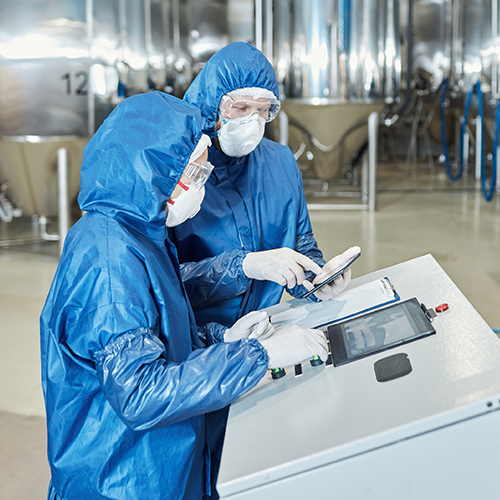 Two technicians wearing blue sterile suits, masks, and gloves operate a touchscreen control panel in a pharmaceutical production facility with stainless steel tanks.