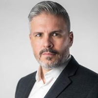 Professional headshot of a man with short gray hair and beard, wearing a dark suit jacket and white shirt, with a serious expression against a light gray background.