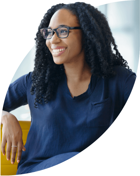 Smiling young woman wearing glasses leans casually against a seat by a large window.