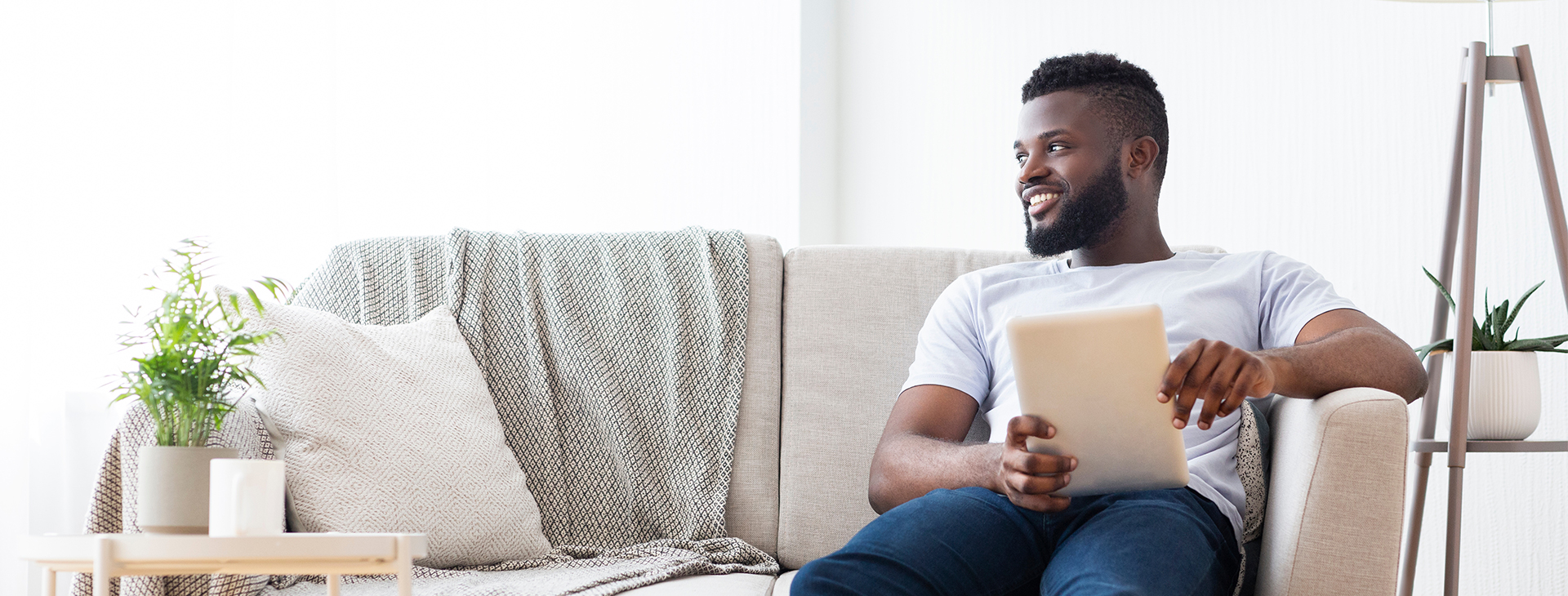 Un homme assis sur son divan, lisant sa tablette et souriant