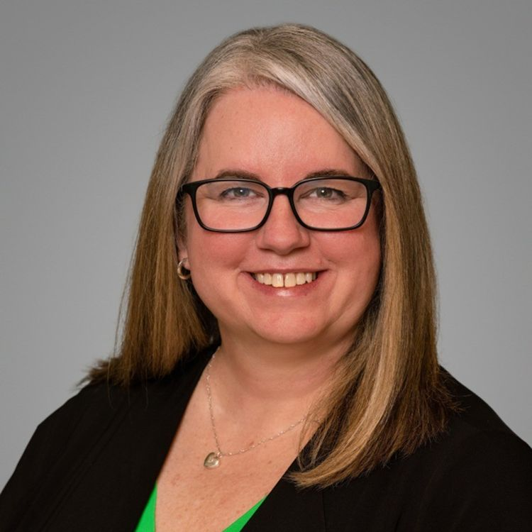 Professional headshot of a smiling woman with long gray-blonde hair and glasses, wearing a black blazer and green top, against a gray background.