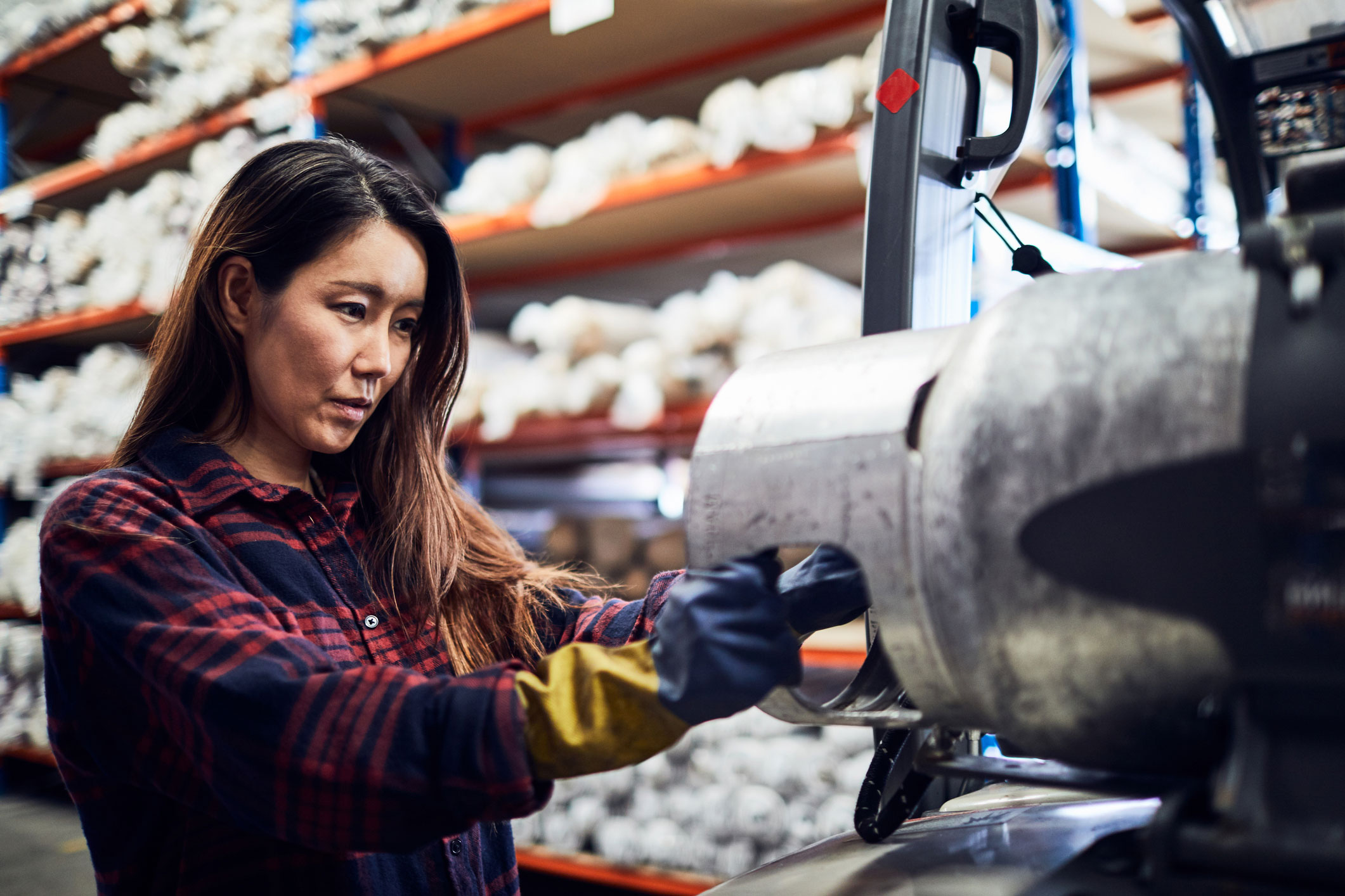 A focused factory worker in a plaid shirt operates an industrial machine amid shelves of material rolls.