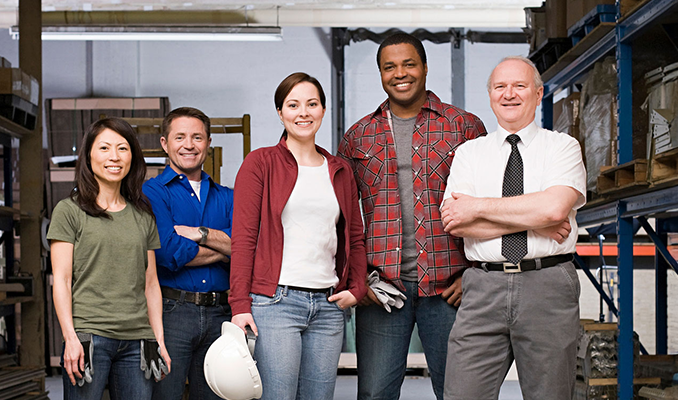 A diverse group of five smiling workers standing together in a warehouse with storage shelves in the background.
