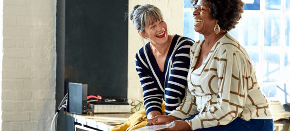 Two mature women sitting on a desk are laughing in conversation.