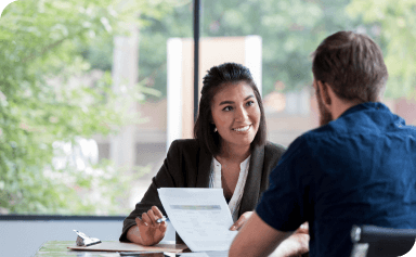 Smiling businesswoman showing document to a male colleague in a meeting room with large windows and trees outside.