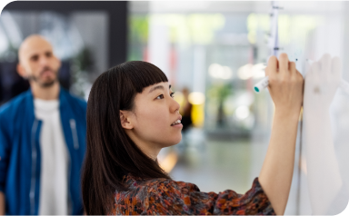 Woman drawing on a whiteboard in an open concept office with two colleagues looking on.