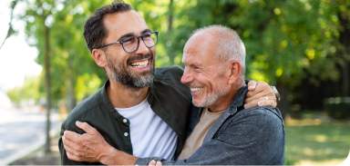 Man and father standing outdoors in a tree-lined street laugh and hug each other.