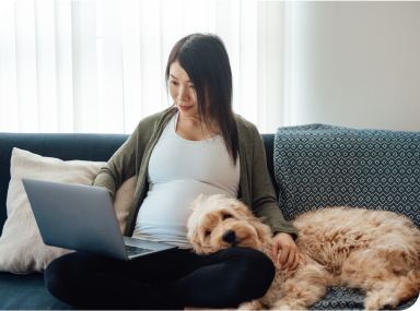 Une femme enceinte est assise sur un canapé, les jambes croisées, et navigue sur un ordinateur portable tandis que son chien repose sa tête sur ses genoux.