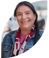 Woman with long braid, patterned scarf and denim jacket smiles at camera with a busy street behind her.